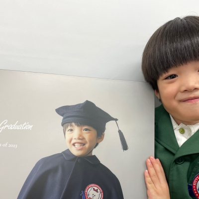 子供が卒業式の衣装を着て、背景には「My First Graduation KIS Class of 2023」と書かれたポスターが写っている記念写真。
