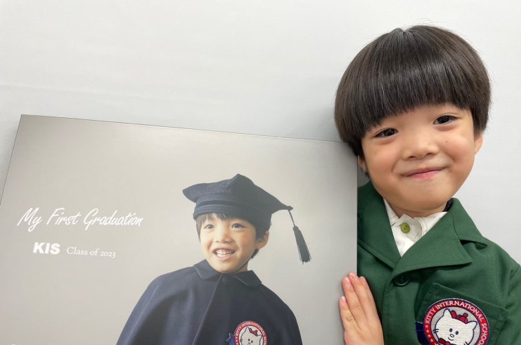 子供が卒業式の衣装を着て、背景には「My First Graduation KIS Class of 2023」と書かれたポスターが写っている記念写真。