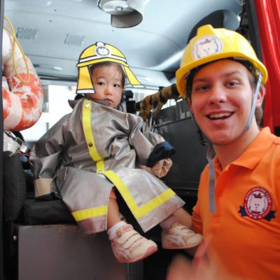 消防士の格好をした子供が消防車の中に座り、隣には笑顔の男性がいる。子供は真剣な表情で消防士の役割を楽しんでいるようだ。